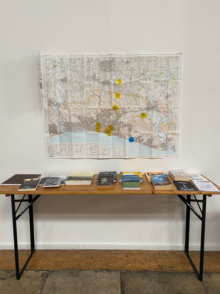 A long wooden table up against a white wall. On the table is a selection of books. Hanging on the wall is a ordnance survey map of Brighton. 