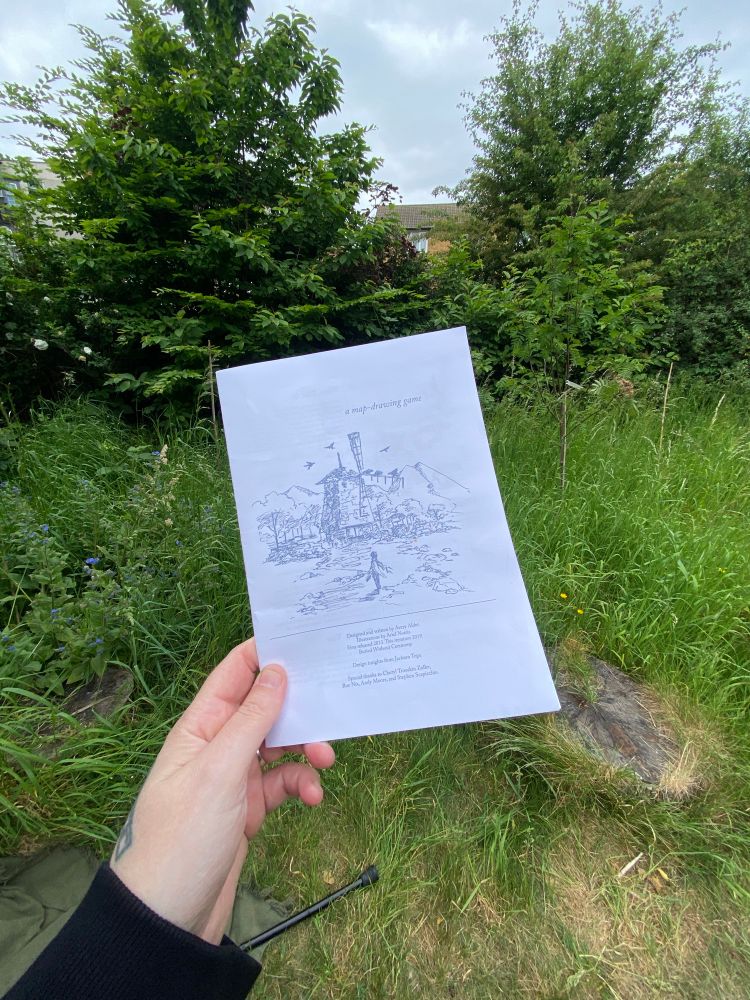 The cover of the TTRPG ‘a quiet year’ held in my hand, with green grass, wildflowers and bushes in the background 