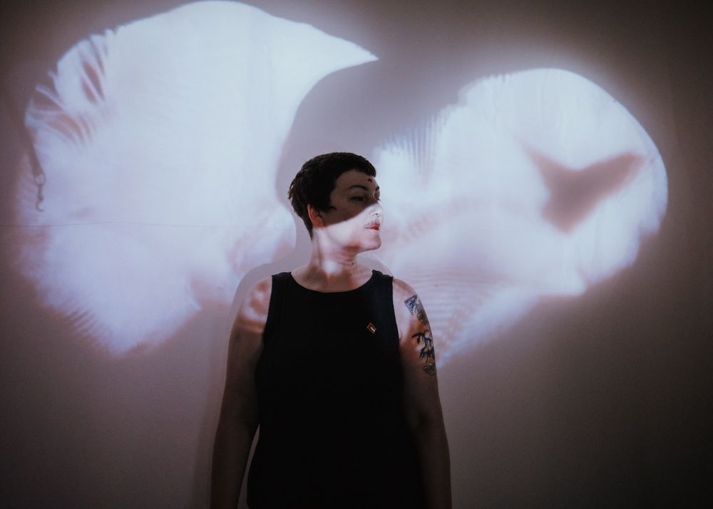 A portrait of me, an artist with dark short hair wearing a black vest top and red lipstick, standing infront of a projection of my work ‘organsongs’. A pink hazy organic shape is projected across my face and body as I turn my head slightly to the right of the scene. 
