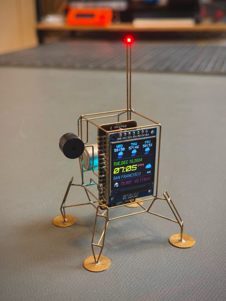 A small lander inspired circuit sculpture with a small screen that is displaying time and weather forecast for the next three days.