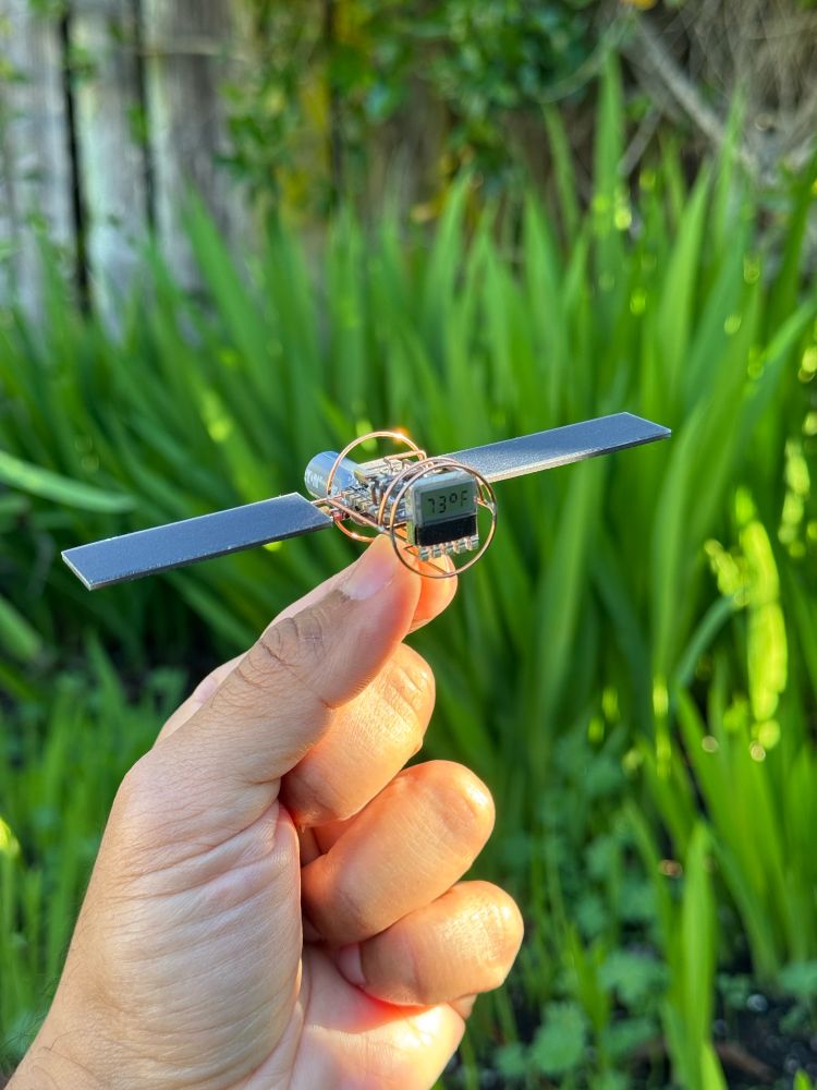 Hand holding a small solar powered circuit sculpture shaped like a satellite. It’s tiny display is showing an ambient temperature reading of 73F. There is green grass in the background. 