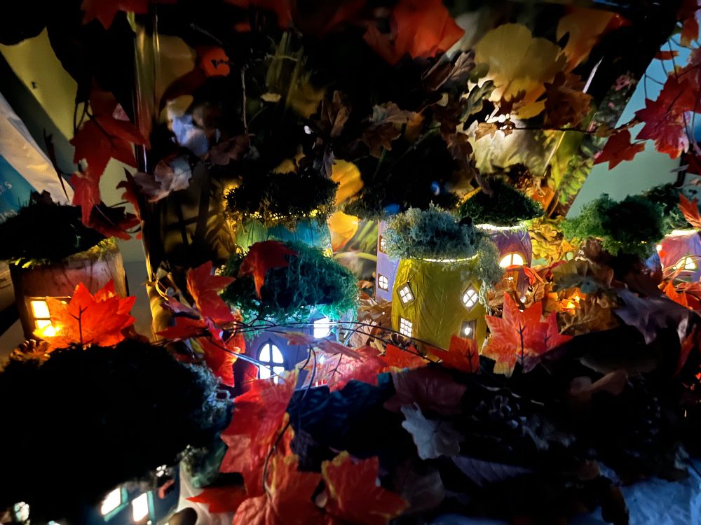 This image shows little fairy houses made out of glass jars, air/dried clay which are the painted bright colours. Each has a glaze painted on it too. They all have fairy lights instead which illuminates the little windows and doors. They’re surrounded by leaves, acorns & chestnuts, making it look like a forest floor. 