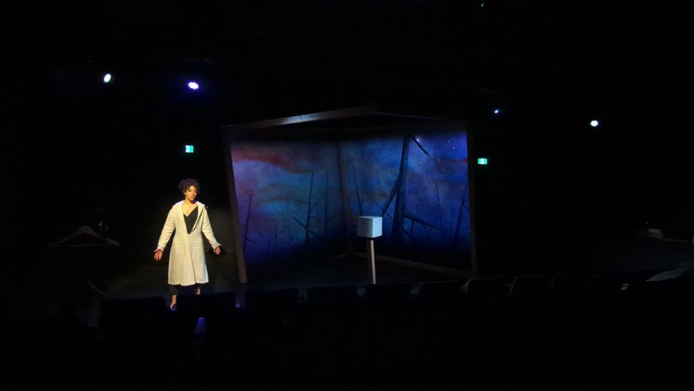 Pandora (played by Jessica b hill) stands next to a cube, the Upstage walls are covered in projections of large spikes.  A white box sits in the middle on a pedestal 