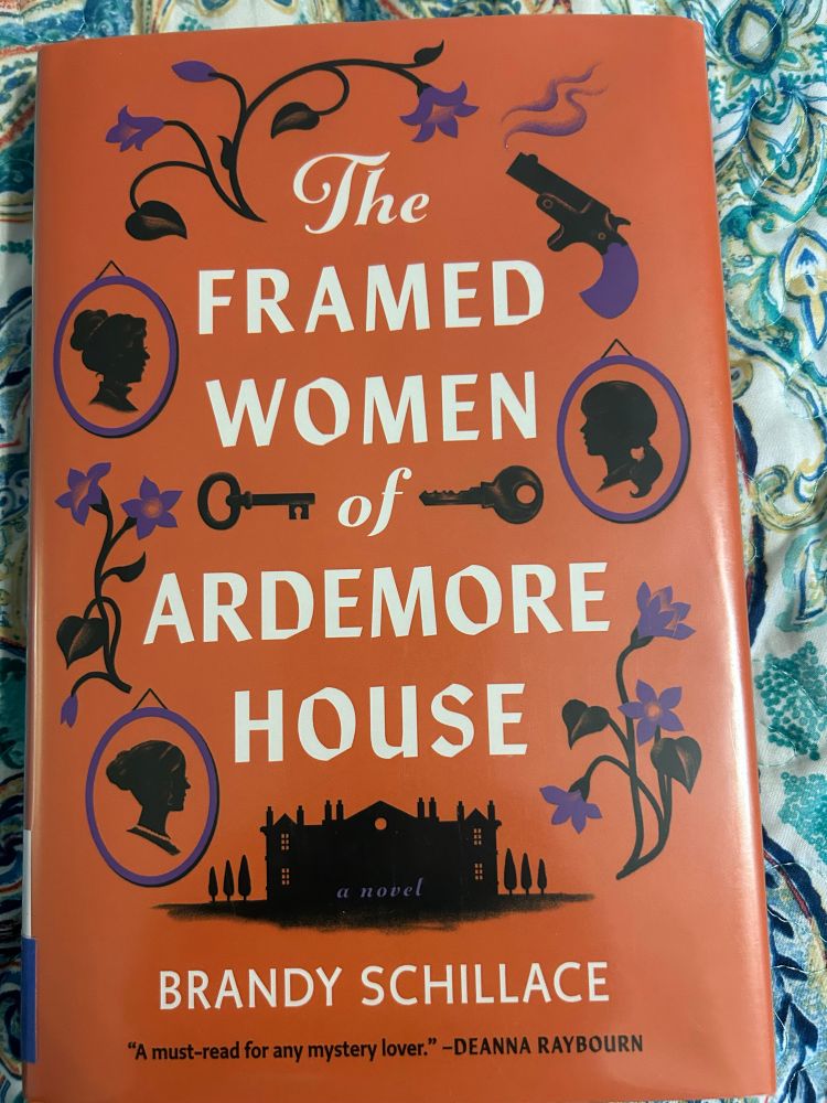 Book cover with orange background, white lettering for title and author: “The Framed Women of Ardmore House Brandy Schillace” designs around the title and author name are in black and purple, with black women’s silhouettes surrounded by purple frames, purple flowers with black stems and leaves, black keys, a black gun with purple handle/grip, and below the title is a black rendering of a manor house with trees on both sides. 