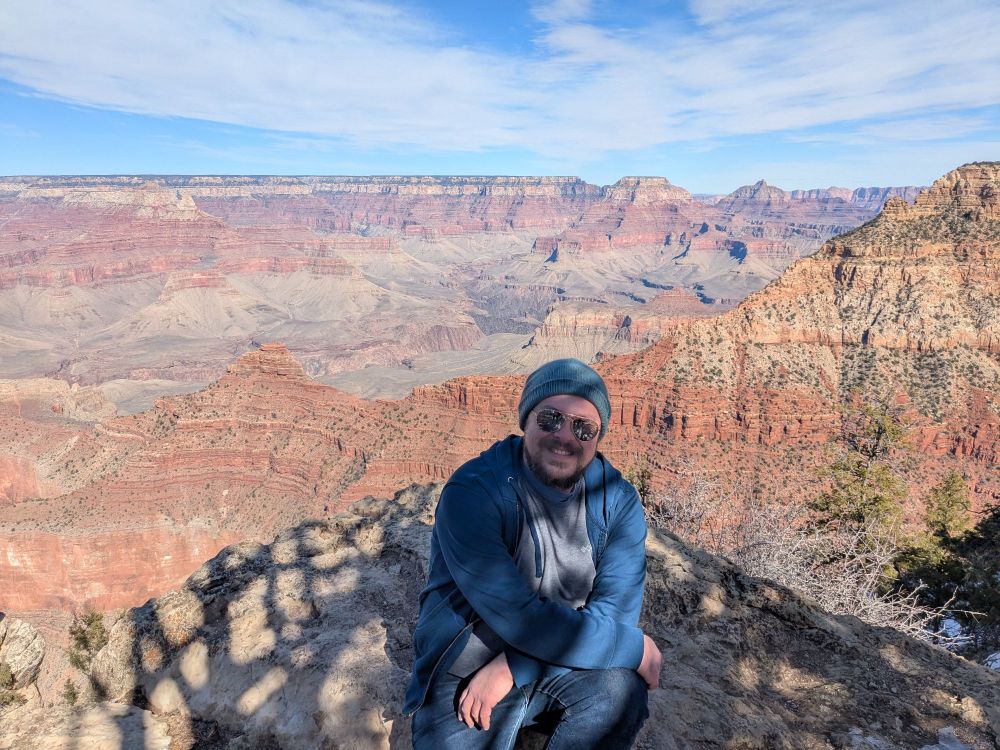 Photo of a man sitting on the edge of a cliff, with the Grand Canyon in the background.
