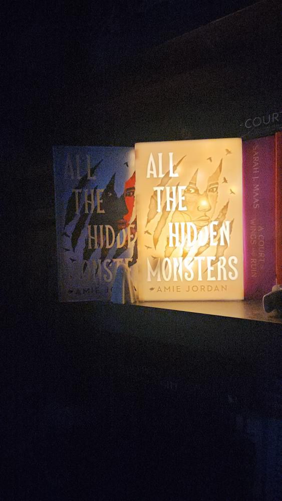 A book shape and size lamp with the cover of ALL THE HIDDEN MONSTERS by Amie Jordan etched into the front 