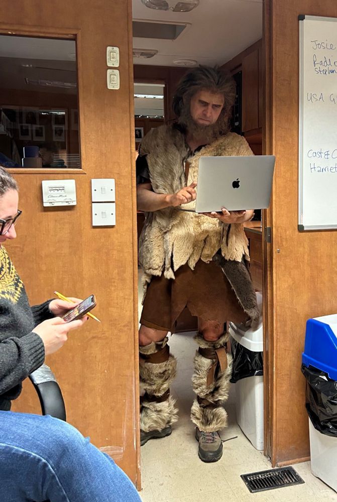 Larry Rickard in most of his costume & makeup for Robin the Ghost (a caveman), in a trailer, looking at a macbook.