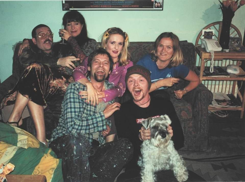 A photo from the millennial sitcom Spaced. 6 people are sharing a tattered greyish 2 person sofa, sitting on each other, arms around each other, smiling. There is a dog and a little bamboo table for the landline phone.