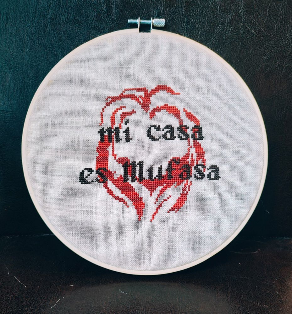 Broderie point de croix dans un tambour en bois. Elle représente une crinière de lion stylisée en dégradé de rouge et le texte "mi casa es mufasa" dessus en marron/noir