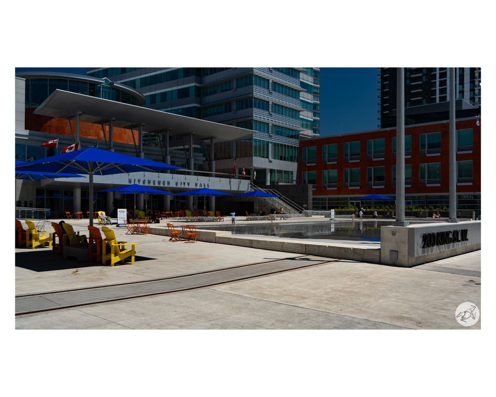 A picture of a splash pad flanked by colourful muskoka chairs & large blue umbrellas that are out front of Kitchener City Hall