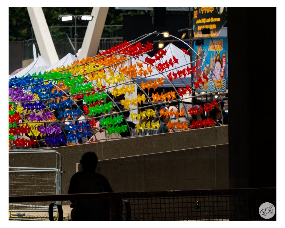 A picture of a festival at Nathan Phillips Square in Toronto, in the background is a tunnel of colourful pin wheels in red, orange, yellow, green, blue & purple. In the foreground is the silhouette of a person sitting in the shade