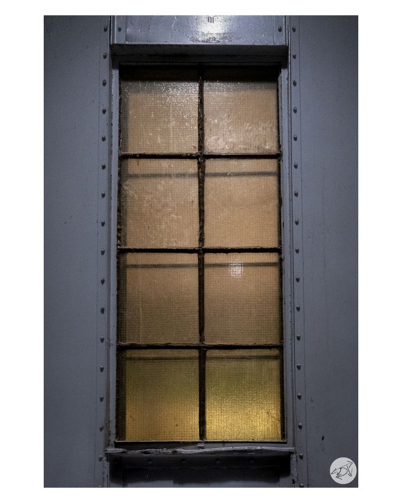 An old window found in Union Station,Toronto. The frames around the hazy glass panes are rusted and there is a warm glow coming through the glass. The window itself is surrounded by the cold steel wall it was intalled into.