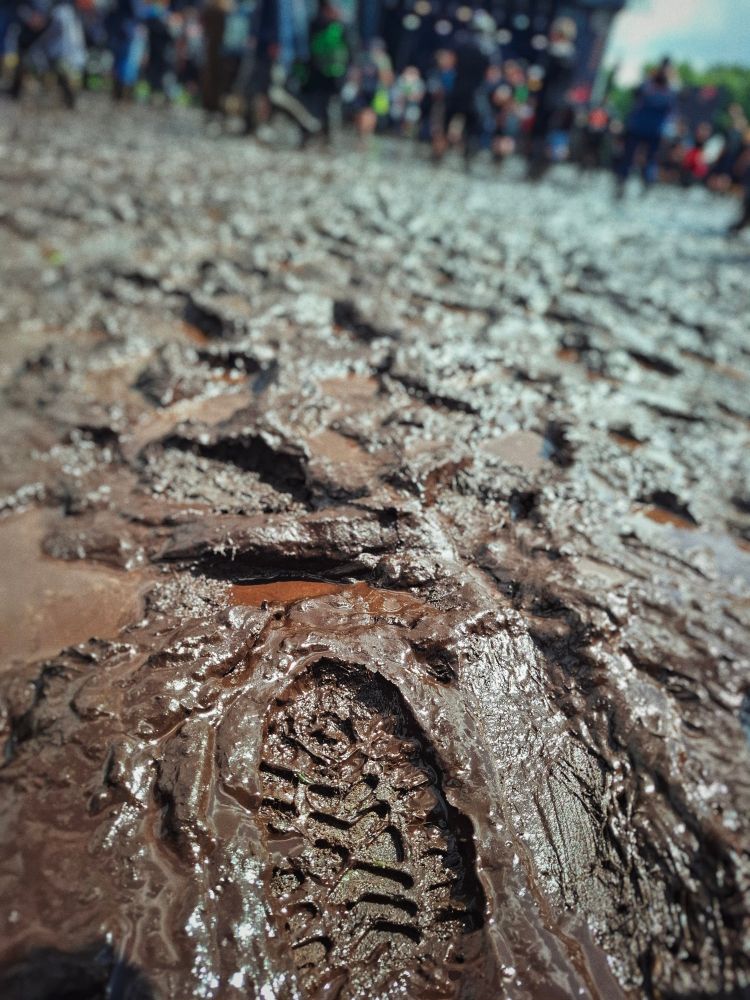A photo of a footprint on the muddy floor, the background is blurred hinting at people standing or walking and a stage with a band playing.