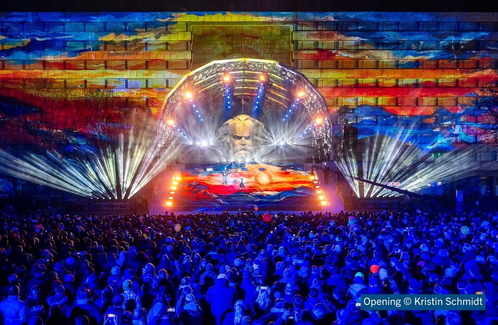 Eine bunt erleuchtete Bühne vor dem Karl-Marx-Monument in Chemnitz mit Scheinwerfern, die Strahlen in alle Richtungen aussenden ist zu sehen. Davor stehen Tausende Menschen in blaues Licht getaucht.