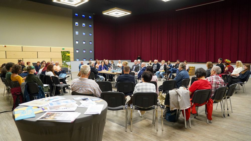 Teilnehmende einer Gesprächsrunde im Luxor Chemnitz sitzen im großen Stuhlkreis um mehrere Tische mit Mikrofonen und Getränken. In der Mitte: Oberbürgermeister Sven Schulze und weitere Vertreter:innen der Stadt im Austausch mit Vereinsmitgliedern. Im Vordergrund ein Stehtisch mit ausliegenden Flyern, im Hintergrund ein roter Bühnenvorhang und eine grau-beleuchtete Wand.