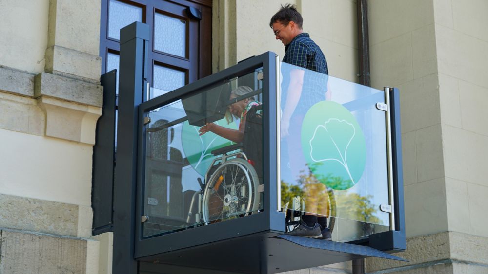 Eine Frau im Rollstuhl und ein Mann nutzen den neuen Plattformlift an der Feierhalle des Krematoriums Chemnitz. Die Glasverkleidung des Lifts ist mit einem grünen Gingko-Blatt-Symbol versehen. Im Hintergrund ist die historische Sandsteinfassade des Gebäudes zu sehen.