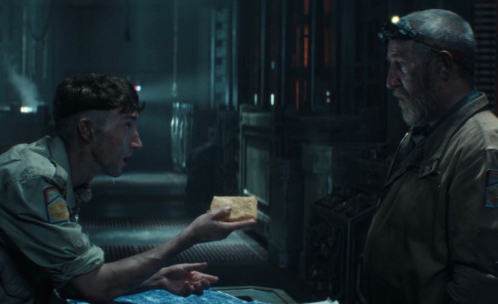 Two men stand opposite each other. One holds a rectangular pastry. 