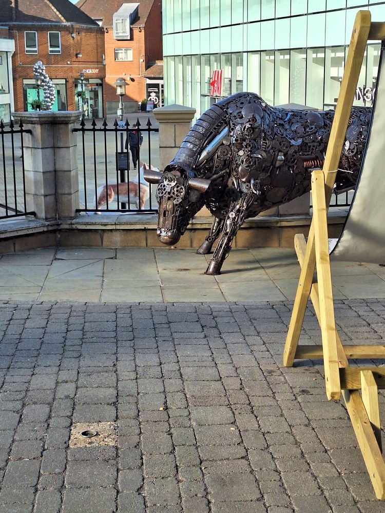 View looking past a double-size fixed deckchair at a cow sculpture made of metal gears and mechanical parts.
 On a lower level behind this is part of an open shopping street, displaying a fiberglass (?) corgi the size of a short-legged horse.