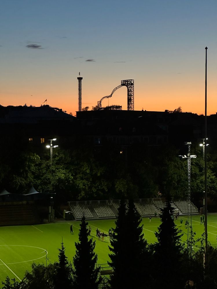 Bragun jalkapallokenttä etualalla ja Linnanmäen laitteet silhuettina taka-alalla iltaruskon aikaan.