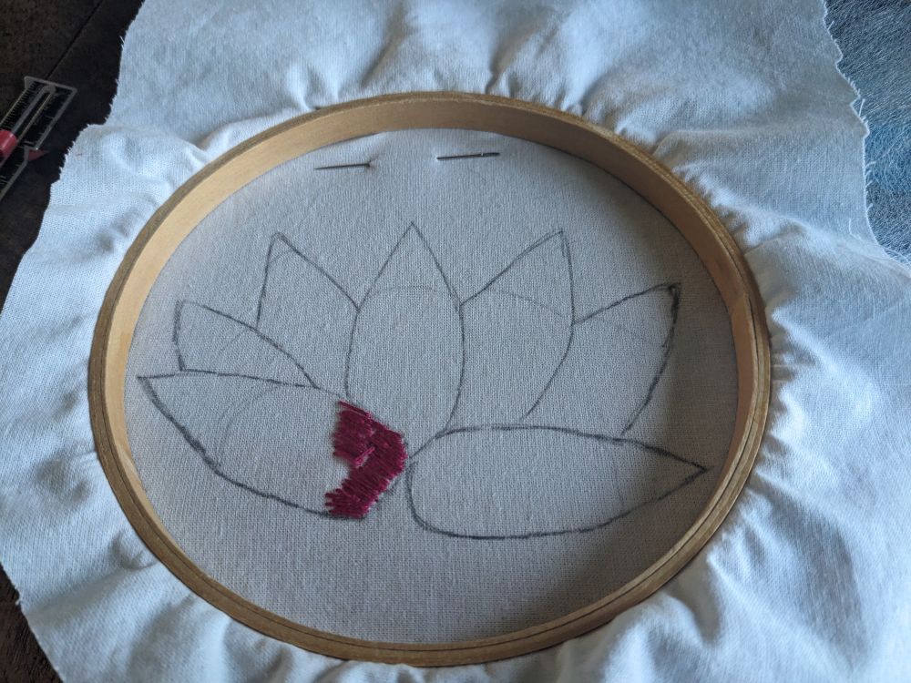 White fabric with a lotus flower line drawing in an embroidery hoop. Some very shoddy initial pink stitches have been started.