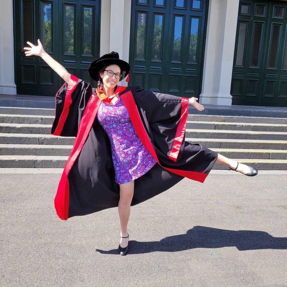 A woman posing in a dancing position. She is wearing a graduation gown and hat, and a dress with a pattern of immune cells 