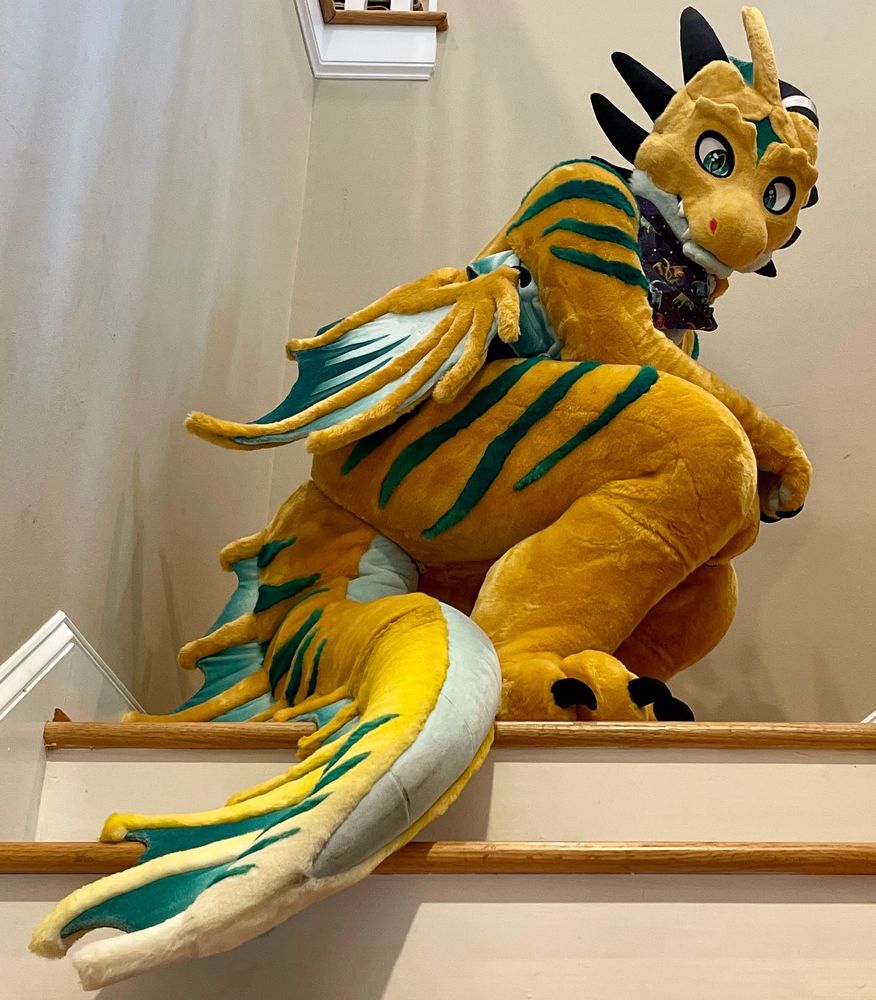 Setrahn the bronze dragon in fursuit form, his tail draped down some stairs yet looking glorious. He is crouched down, looking side eye at the camera.