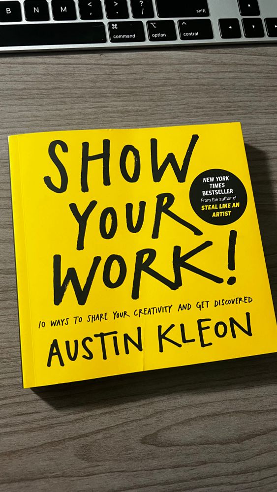 Bright yellow cover of the book Show Your Work! by Austin Kleon, placed on a wooden desk in front of a MacBook keyboard.