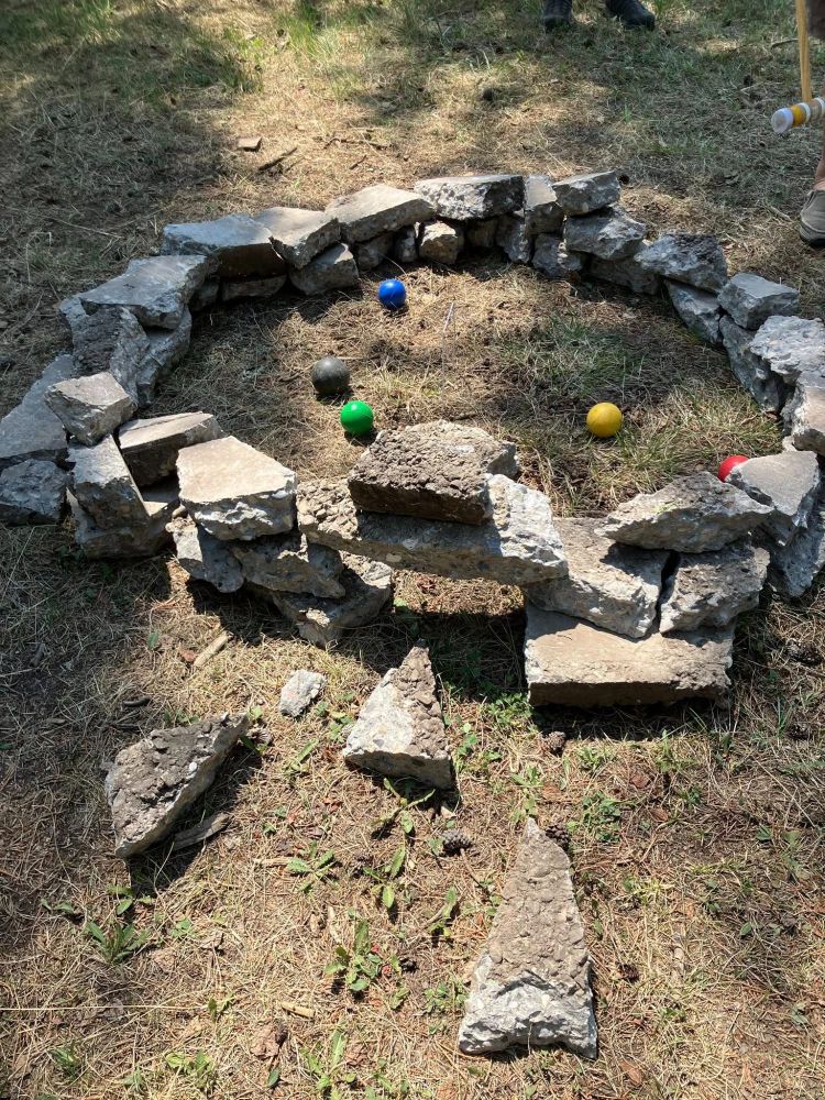 A ring of jagged concrete blocks with 5 coloured croquet balls surrounding a central wicket.