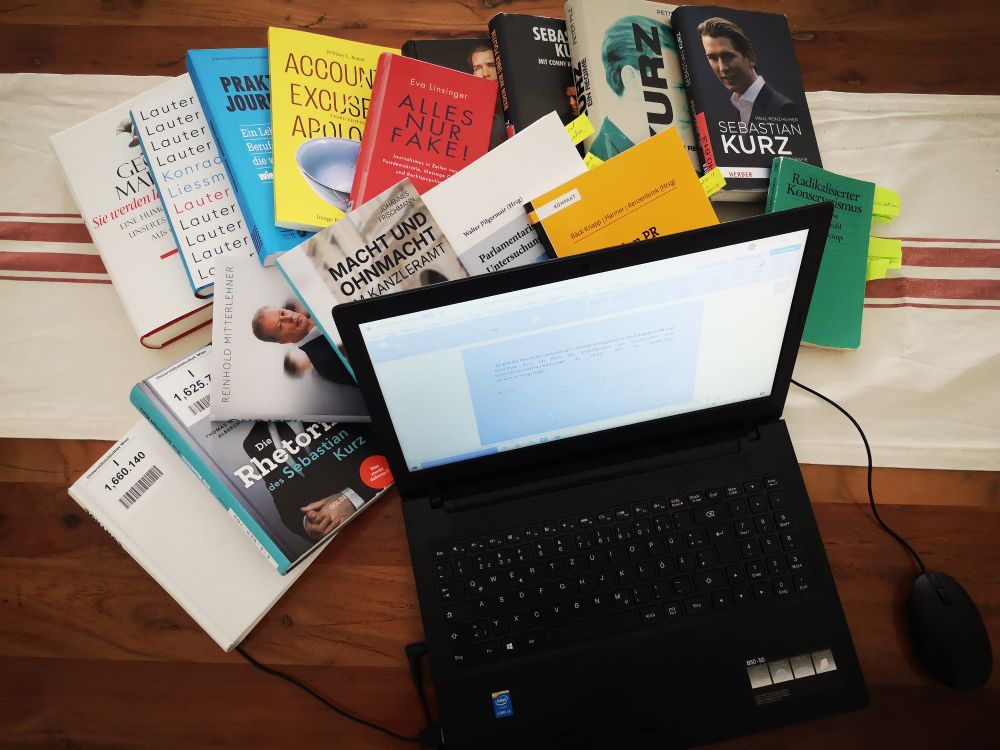 Ein Holztisch aus Vogelperspektive. Darauf steht ein Laptop mit einem offenen Dokument. Hinter dem Laptop liegt ein großer Haufen Bücher zu Sebastien Kurz, österreichischer Politik, Litigation PR und Fake News.
