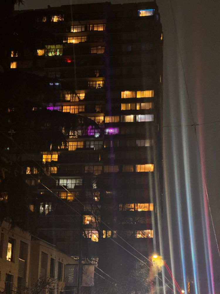 Car Headlights streaming up into the night fog in the colors of the trans flag against the backdrop of an amber and purple lit condo 