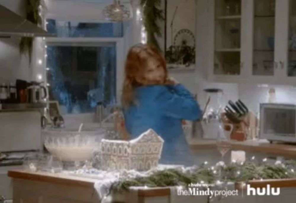 a woman in a kitchen with a gingerbread house on the counter