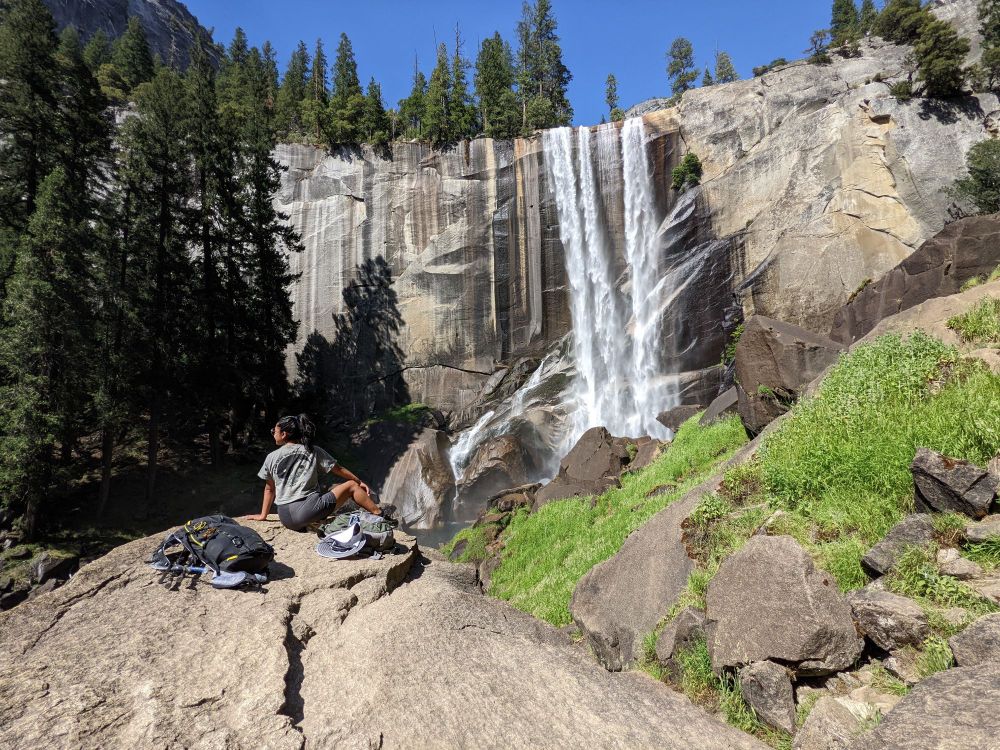 A person sits on a large granite rock, looking towards Vernal Fall in Yosemite National Park. The waterfall powerfully cascades down a sheer granite cliff, surrounded by pine trees on the left and a green, grassy slope on the right. The person's backpack and shoes are next to them on the rock.