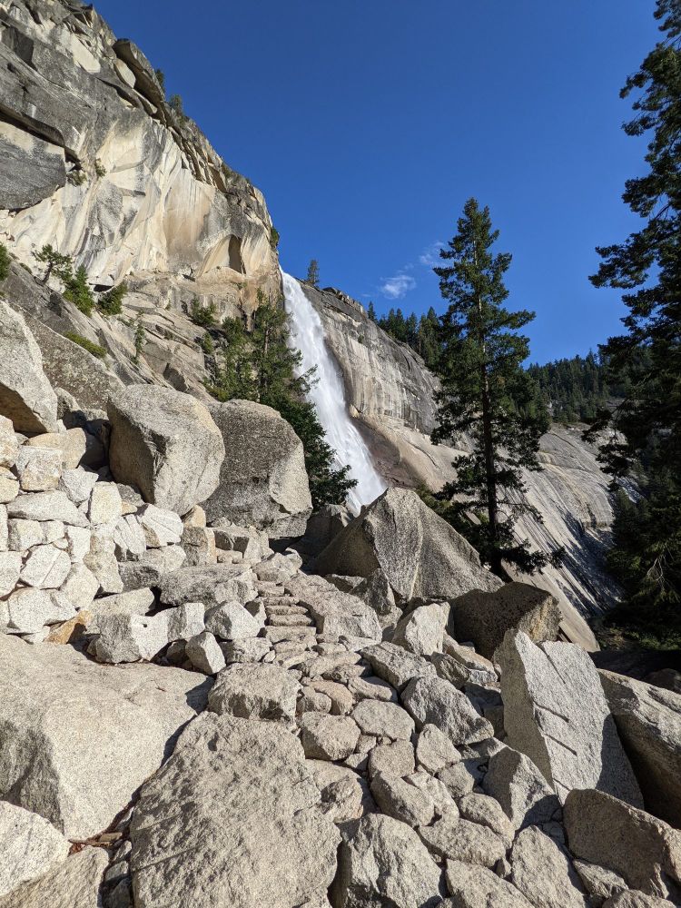 A view from the Mist Trail shows a rocky path winding up towards Vernal Fall. The waterfall is visible in the background, flowing down a smooth granite cliff. The foreground is filled with large boulders and small trees under a clear blue sky.