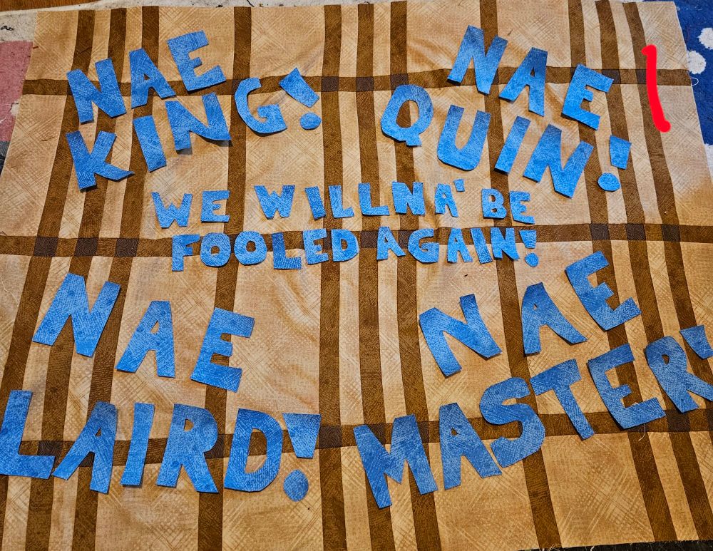 All of these are Tan and brown fabric pieced together to look like tartan. Laying in it are blue block letters that spell out "Nae King! Nae quin! Nae Laird! Nae master! We willna' be fooled again!"

For 1: the Nae phrases are one in each corner with we willna'... In the center