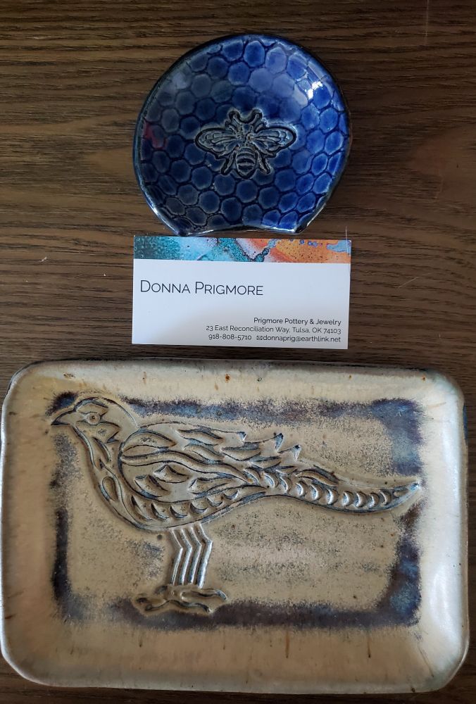 Two small pottery pieces on a table top.
The top is a small, round, cobalt blue chopsticks rest. It has impressions of honeycomb and a bee.
The bottom is a tray, 3x5, with a high-relief impression of a pheasant. It's sand colored, with blue accents.
The artist's business card is placed between. Donna Prigmore
Prigmore Pottery & Jewelry 
23 East Reconciliation Way
Tulsa, OK 74103. 918-808-5710
Donnaprig@earthlink.net