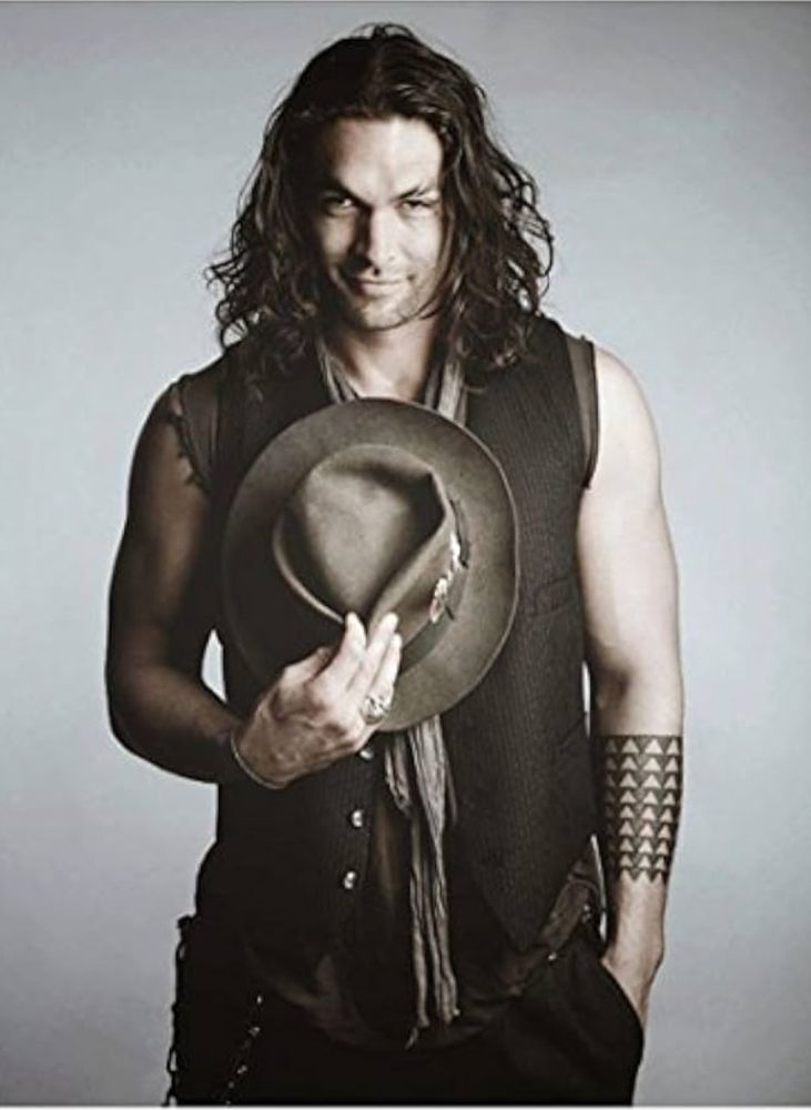 The actor Jason Momoa wearing a black vest, holding a hat, grinning at the camera. 