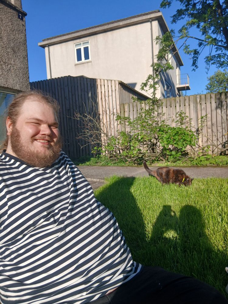 Máni sætt og brosandi í sólinni á grasbletti með Skottu tortie kisu að borða gras í bakgrunni
