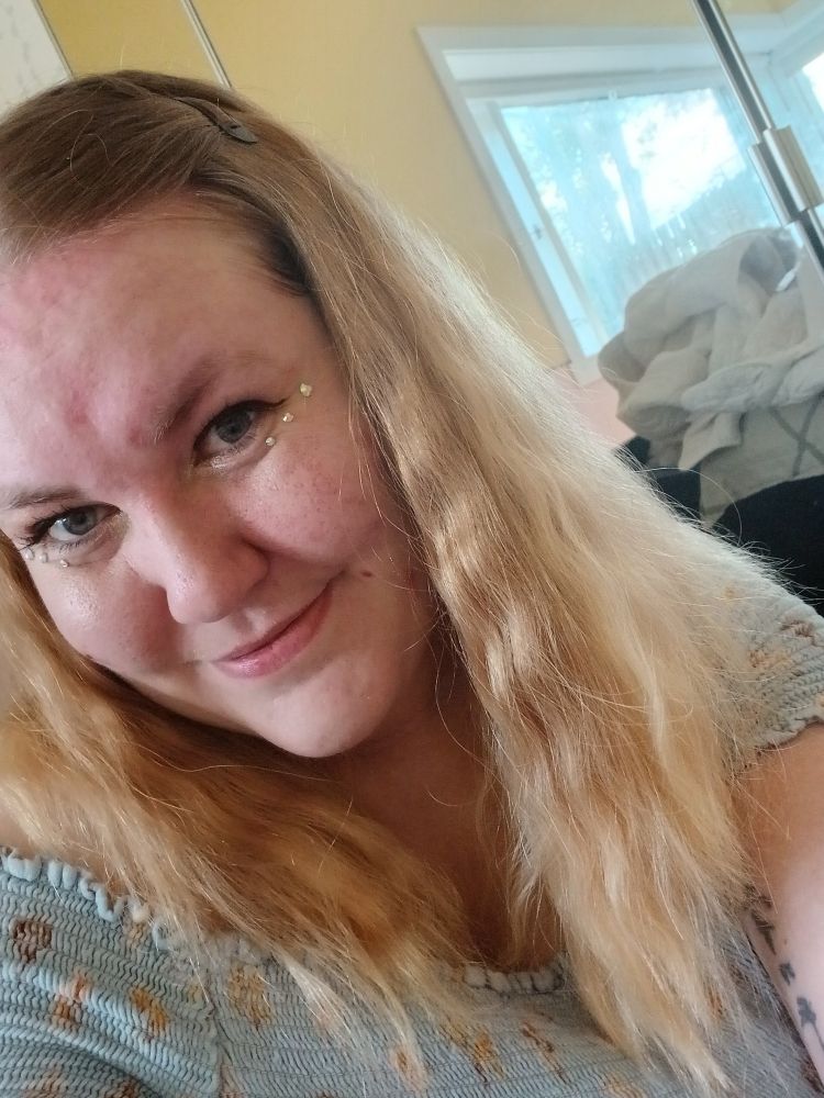 Sjálfa af mér með ljóst liðað hár í spennu, vængjaðan eyeliner, óljósa glimmeraugnskugga, og þrjá litla gimsteina undir hvoru auga. Ég brosi, í bláum riffluðum blómakjól. Fyrir aftan sést þvottur í kuðli í speglinum.