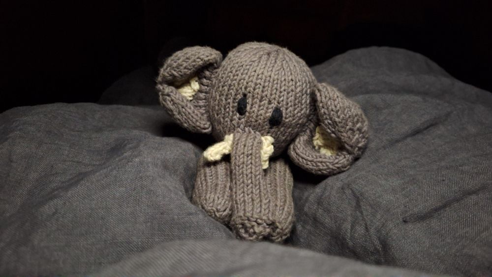 Ein kleiner grauer, gestrickter und äußerst fotogener Plüschelefant, der es sich auf einer grauen Leinendecke gemütlich gemacht hat
