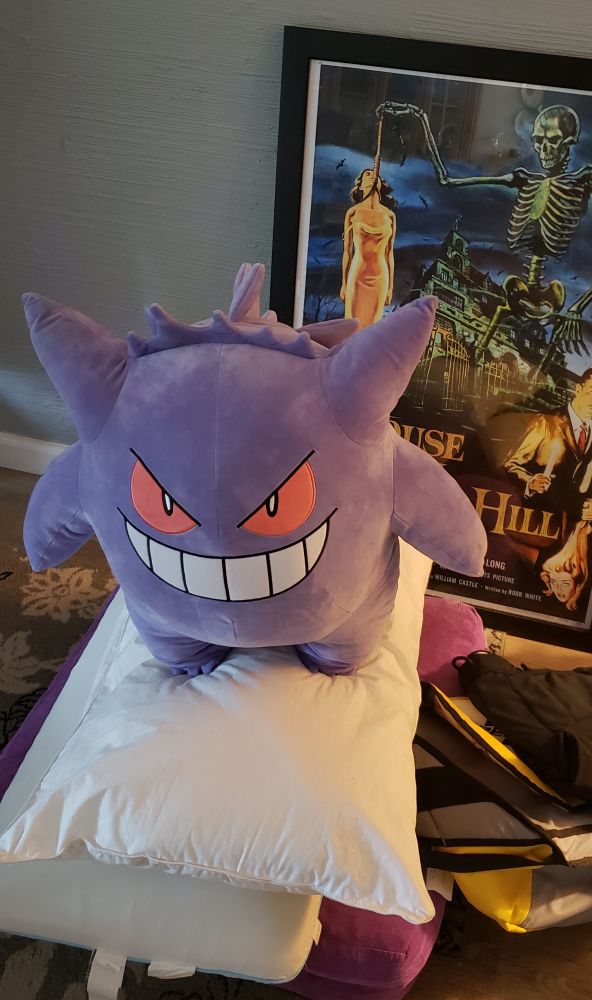 a Gengar plush sits on top of some pillows, a bag, and un-hung paintings