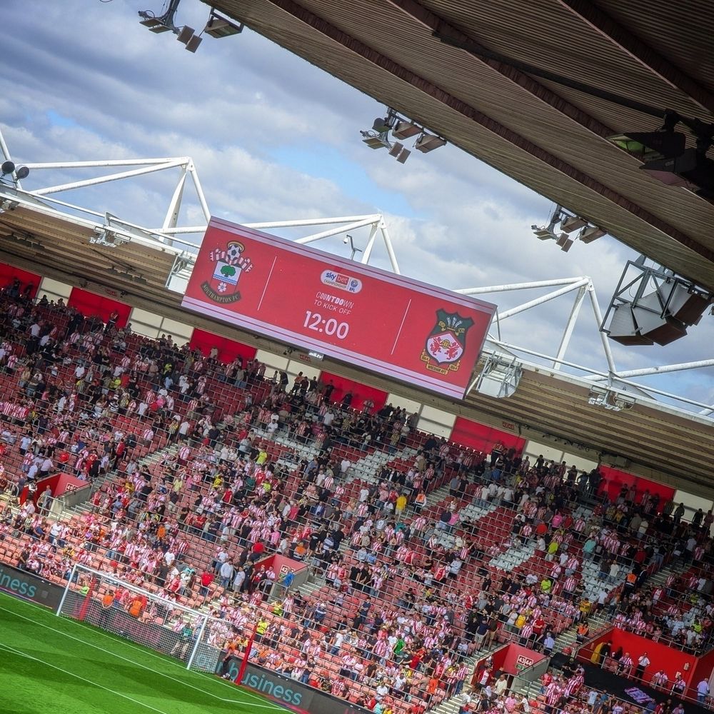 A view from the stands of St. Mary’s stadium, looking towards a large scoreboard showing pre-match info for the match between Southampton FC and Wrexham AFC. The stands are filled with spectators, many wearing red and white.