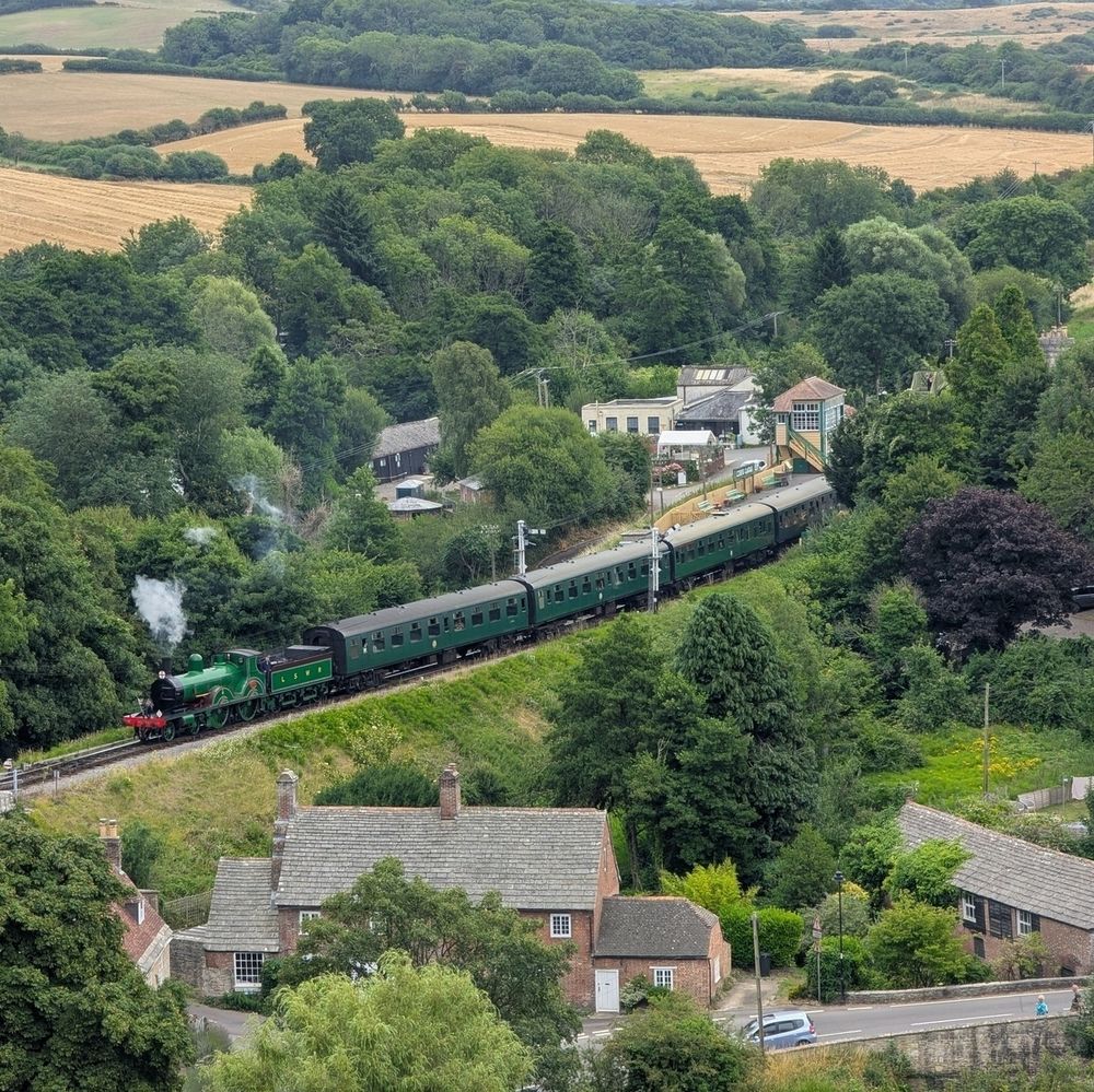 An elevated, aerial view of a green steam train with several passenger carriages traveling on tracks through a green, wooded landscape. The train emits white smoke from its front. In the background, golden fields stretch into the distance, and in the foreground, parts of residential buildings with grey rooves are visible.