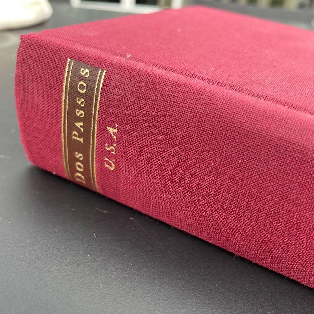 a red hardcover copy of the U.S.A. Trilogy by Dos Passos laying at an angle on a black table