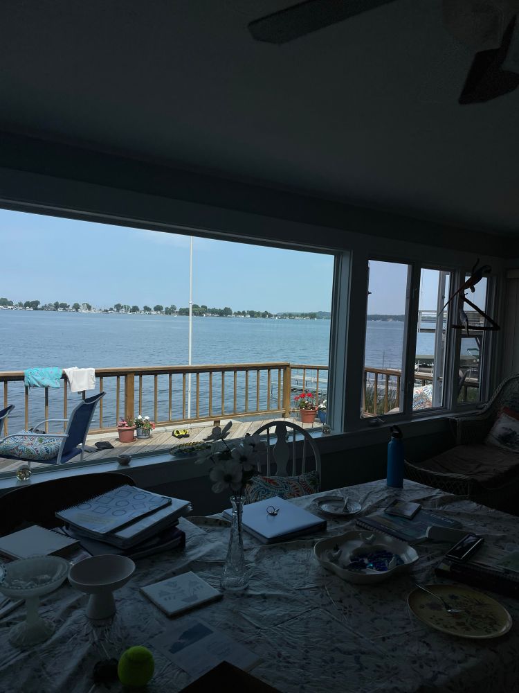view out a window with a table with books and binders in the foreground. wooden deck outside a window with a water bay beyond it 