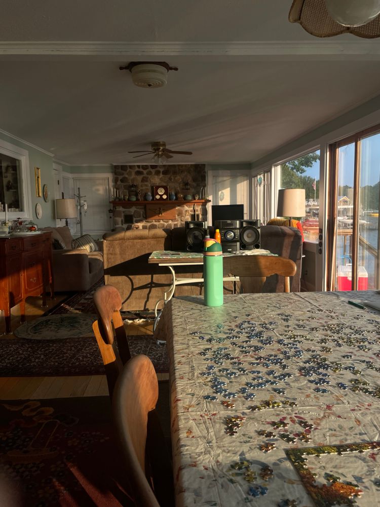 room with a table with puzzle pieces in the foreground with a green owala on it. wooden chairs around it. in the background are couches and a fireplace. sunlight coming in from the right side at an angle that lights up half the room in golden morning light 