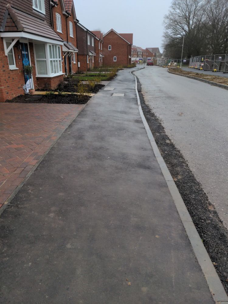 A pavement through a new development with houses on the left hand side and a road on the right hand side.