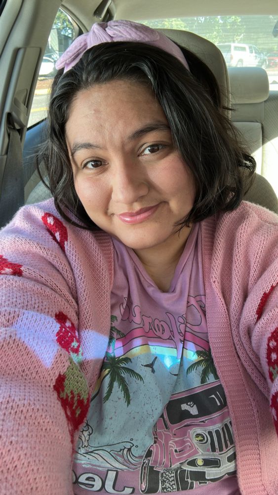 Me wearing a light pink California shirt with a pink Jeep on it with a rainbow and palm tree and beach behind it, and a light pink sweater with strawberries, and a pastel purple headband with a bow on it. I’m smiling at the camera and my wavy brown hair is just slightly messy and goes up to my chin.