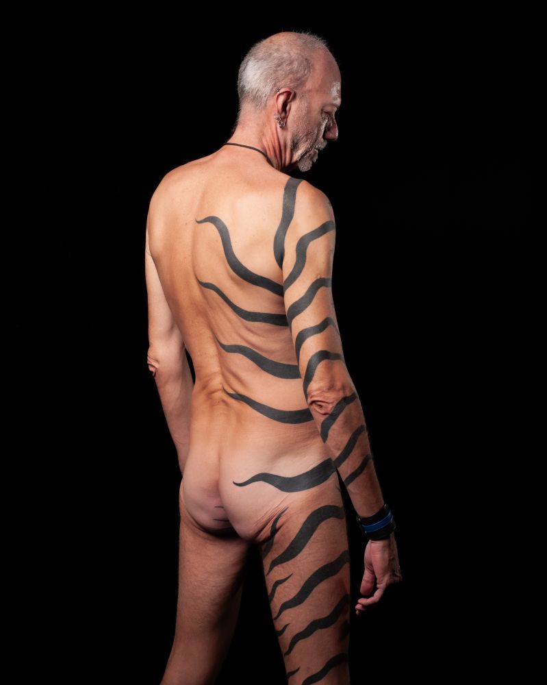 Cute older leather bottom guy, standing, showing off the stark rear and side details of his half body tiger stripes tattoo.