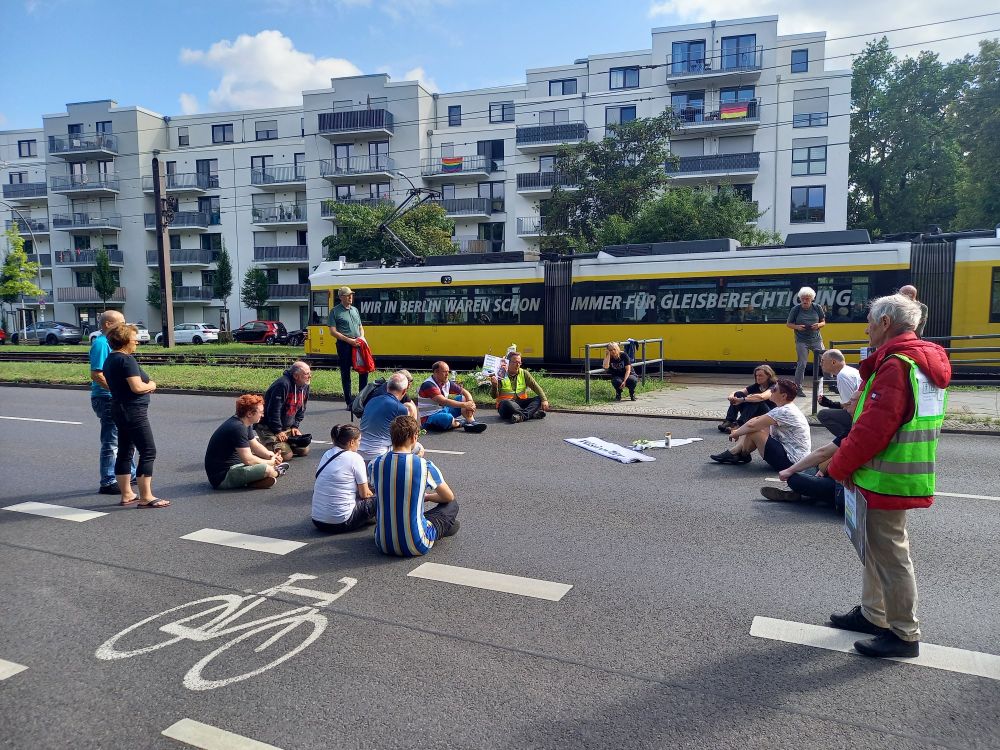 Mahnwache mit verschiedenen Personen die für eine Schweigeminute auf der Fahrbahn sitzen. Im Hintergrund fährt eine Tram.