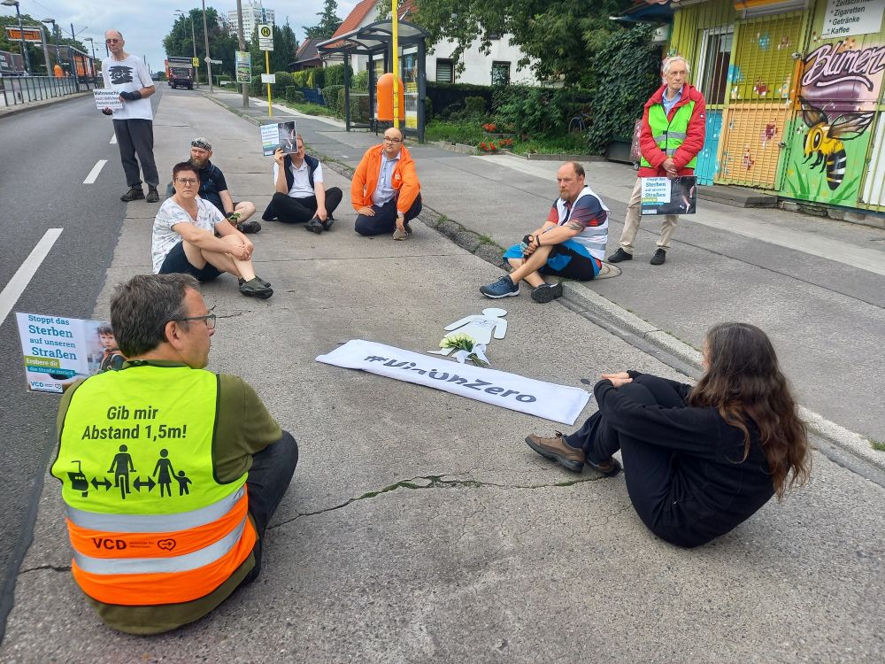 Mahnwache mit verschiedenen Personen die für eine Schweigeminute auf der Fahrbahn an einer Busstation sitzen.